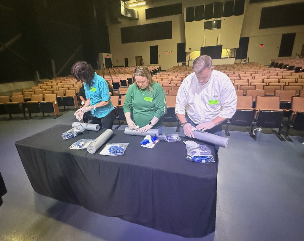 Three people standing by a table with a black tablecloth. They are wearing gloves and learning how to stop the bleed (first aid).