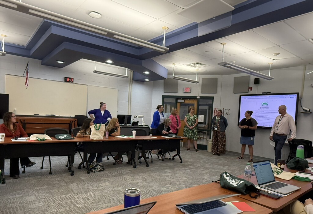 7 people standing in front of a room with others sitting at tables. They are introducing themselves to the new employees during new employee orientation.