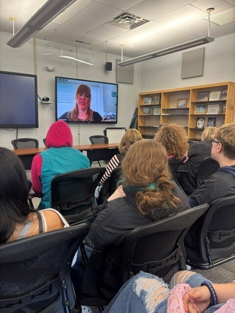 Students listening to the presenter who is virtual. The students are sitting in chairs looking at a large screen with the presenter.