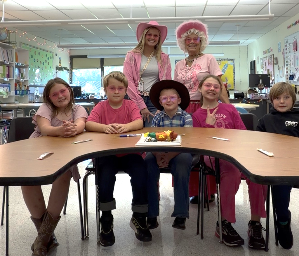 Two staff members dressed in pink standing behind 5 students sitting at a table smiling for the camera