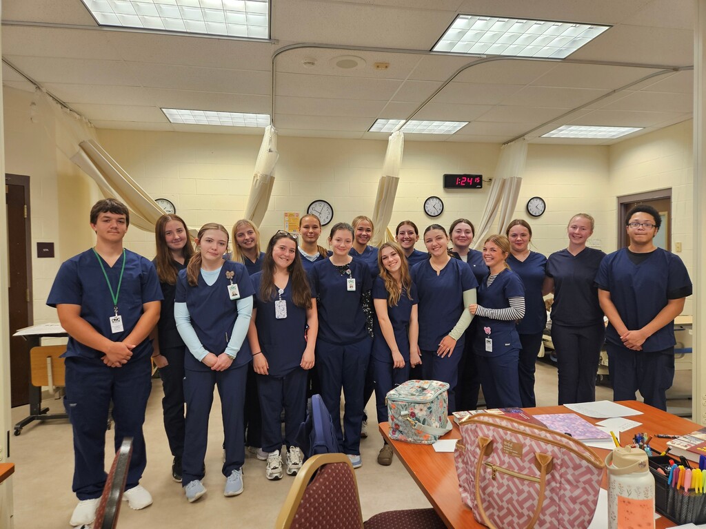 16 students in blue medical scrubs posing for a group picture.