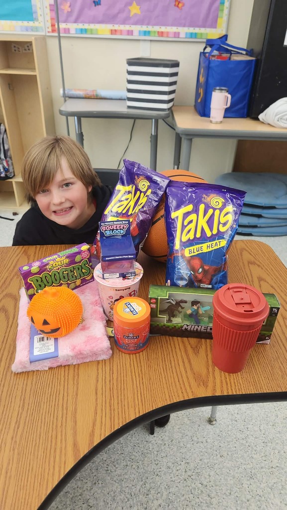 A student sitting at a table with multiple items from the auction including two bags of Takis, candy, a jack-o-lantern toy, and a cup.