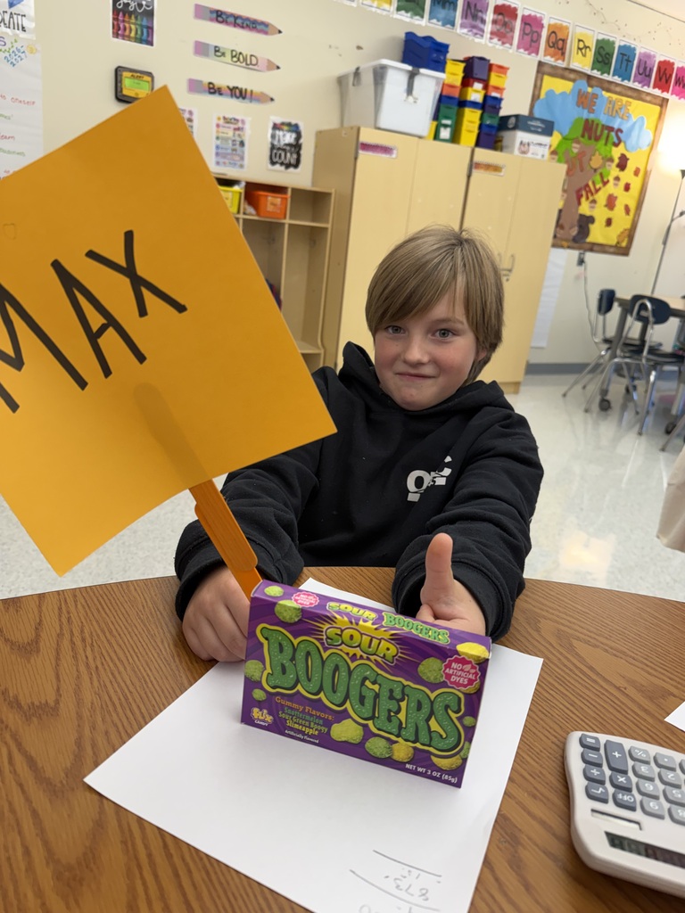Student sitting at a table with a box of candy holding a yellow sign that says MAX