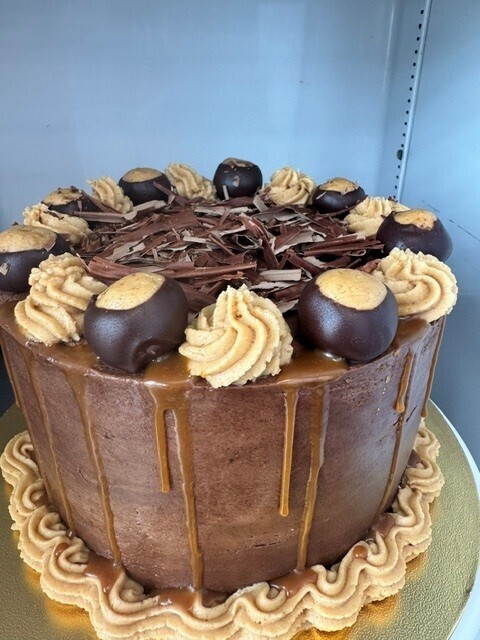 the cake is decorated with brown frosting with a caramel drip. There are chocolate shards on top with piped rosettes in beige frosting and chocolate and peanut butter bon bons in between the rosettes.