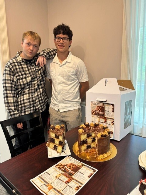 Two students standing behind a table with the cake on it. The cake has a large slice cut out and on a plate while the rest of the cake is on a cake plate.