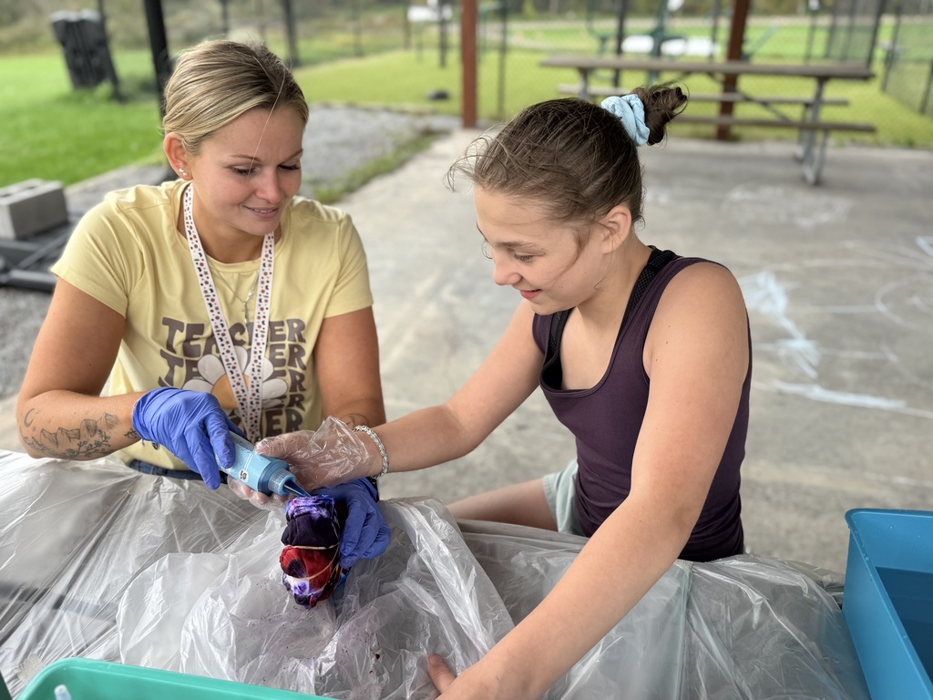 A staff member and a student, both wearing gloves, are working with paint to put on a shirt