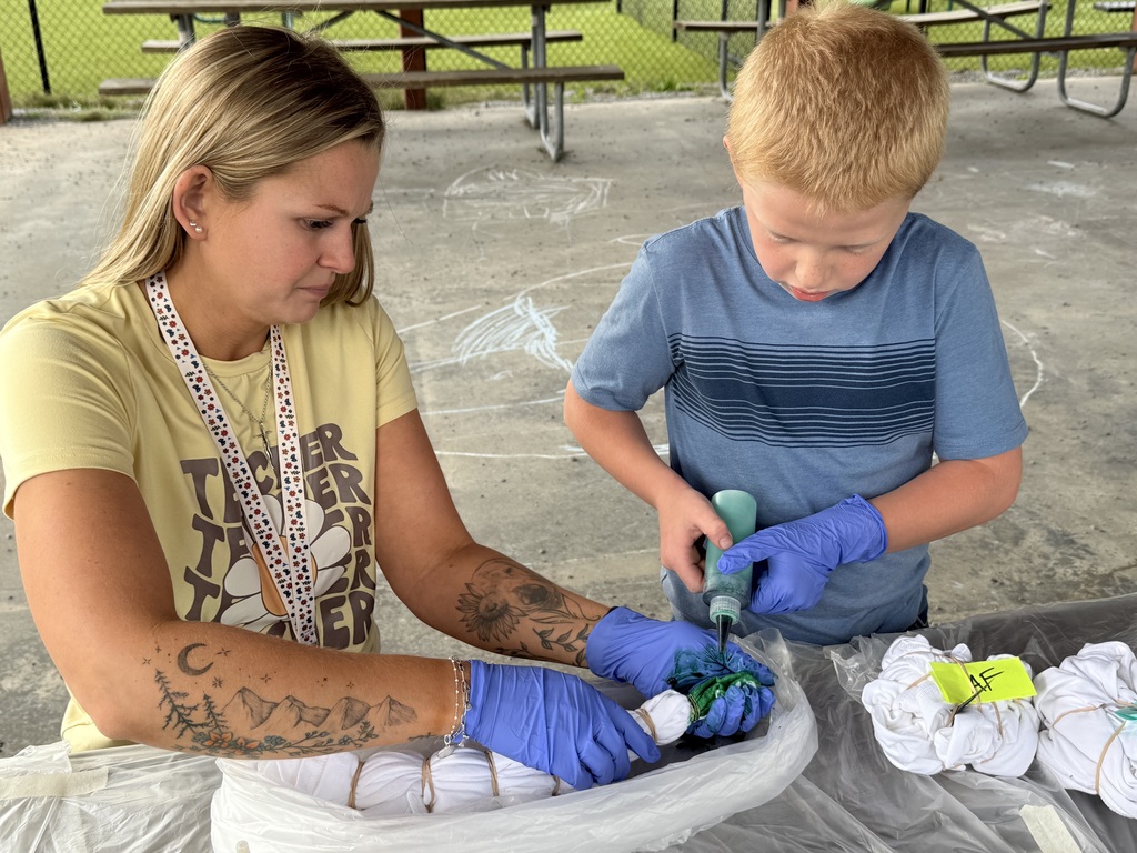 A staff member and a student, both wearing gloves, are sitting at a table outside using green paint to put on a shirt