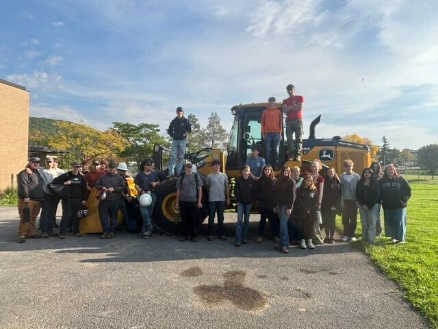 About 30 students standing in front of and on a large yellow tractor outside.