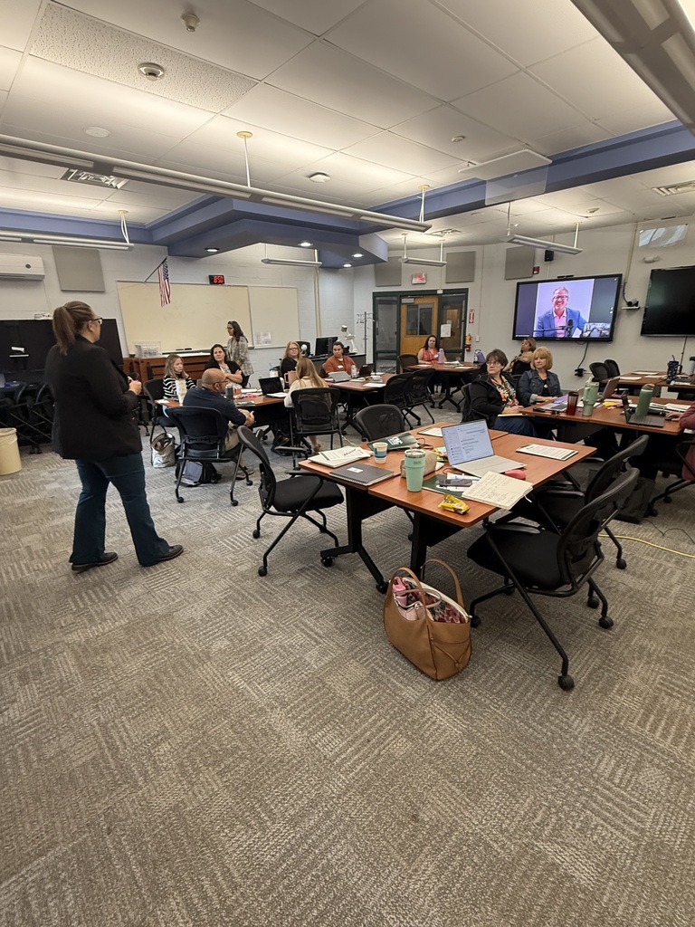 A presenter standing in the from of the room speaking to the participants who are sitting at smaller groups of tables