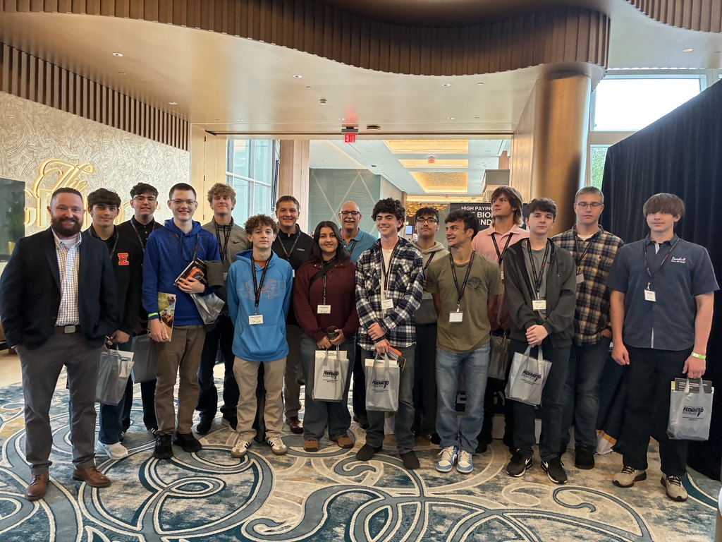 13 students, the instructor, and two adults are standing in a hallway posing for a group picture. The students have lanyards on their necks and are holding bags.