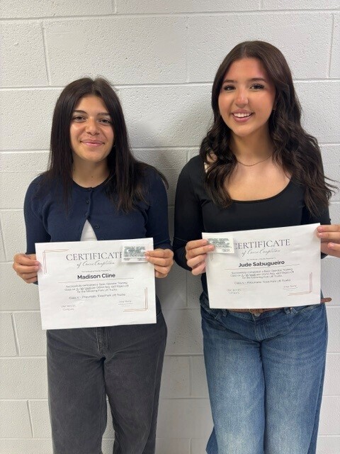 Madison and Jude standing against a white brick wall holding their forklift certificates.