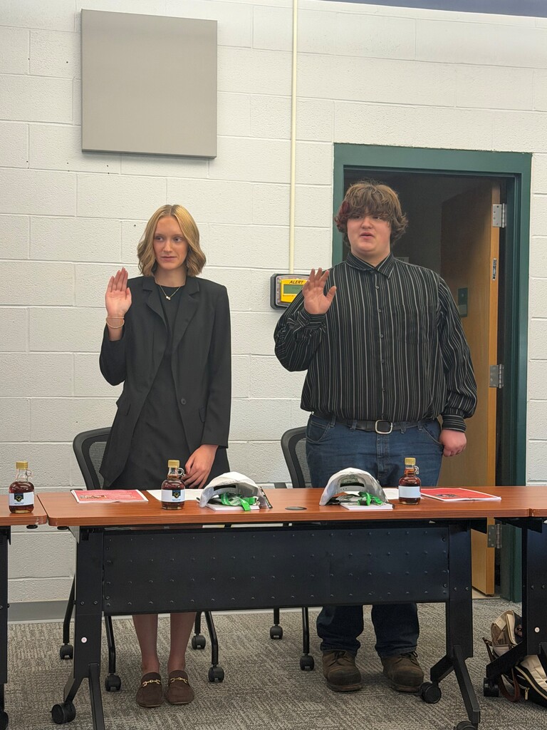 New student board members, Lily and Ashton, standing behind desks with their right hands raised being sworn into their roles.