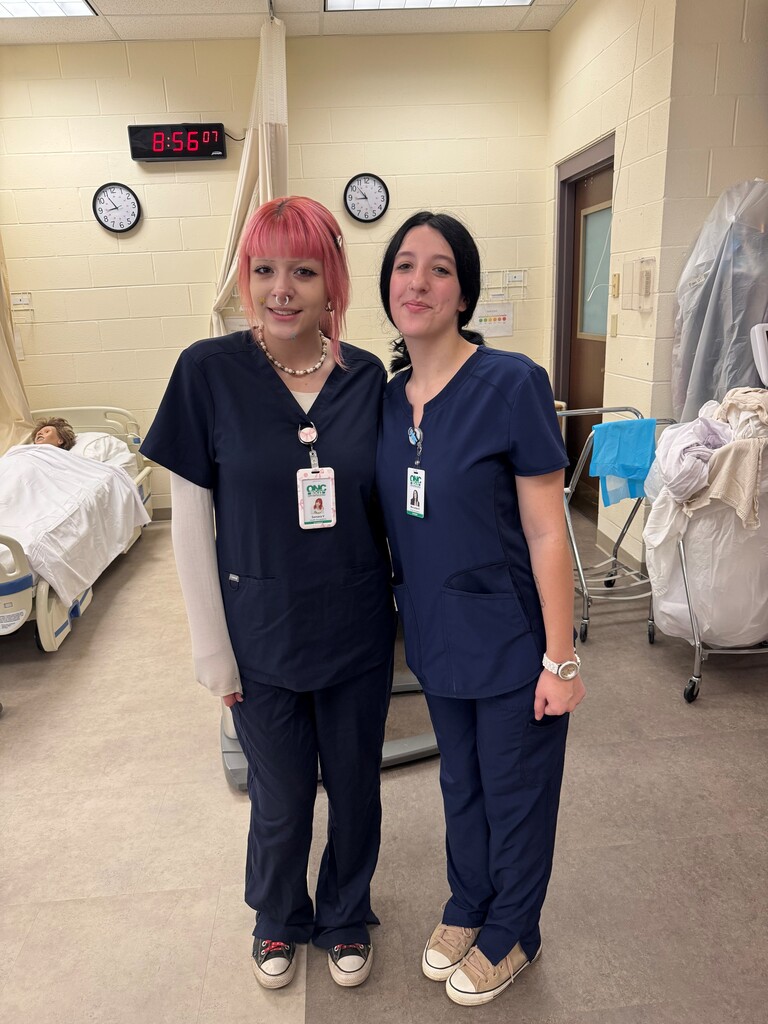 Two students in blue medical scrubs posing for a picture in a clinical teaching room with a mannequin laying in a bed behind them.