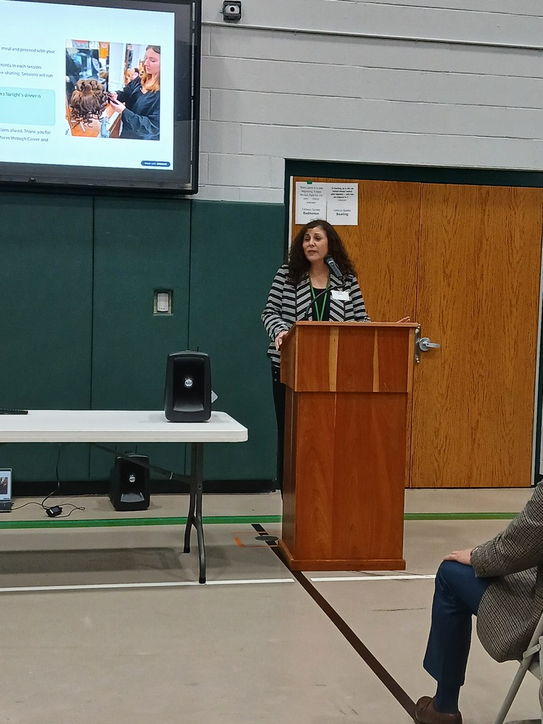 Presenter standing at a podium with a large screen behind them with a picture of a cosmetology student doing hair. The presentation is happening in a gymnasium.