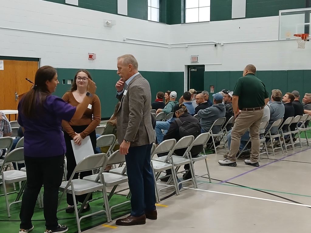 Picture of three people standing and talking in a gymnasium with metal folding chairs lined up for guests. There are other people sitting in chairs.