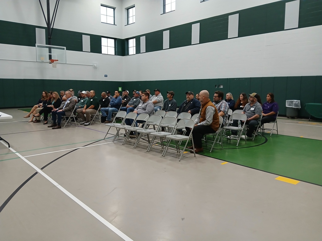 A wider angle of the guests sitting in chairs in the gymnasium listening to the presenter.