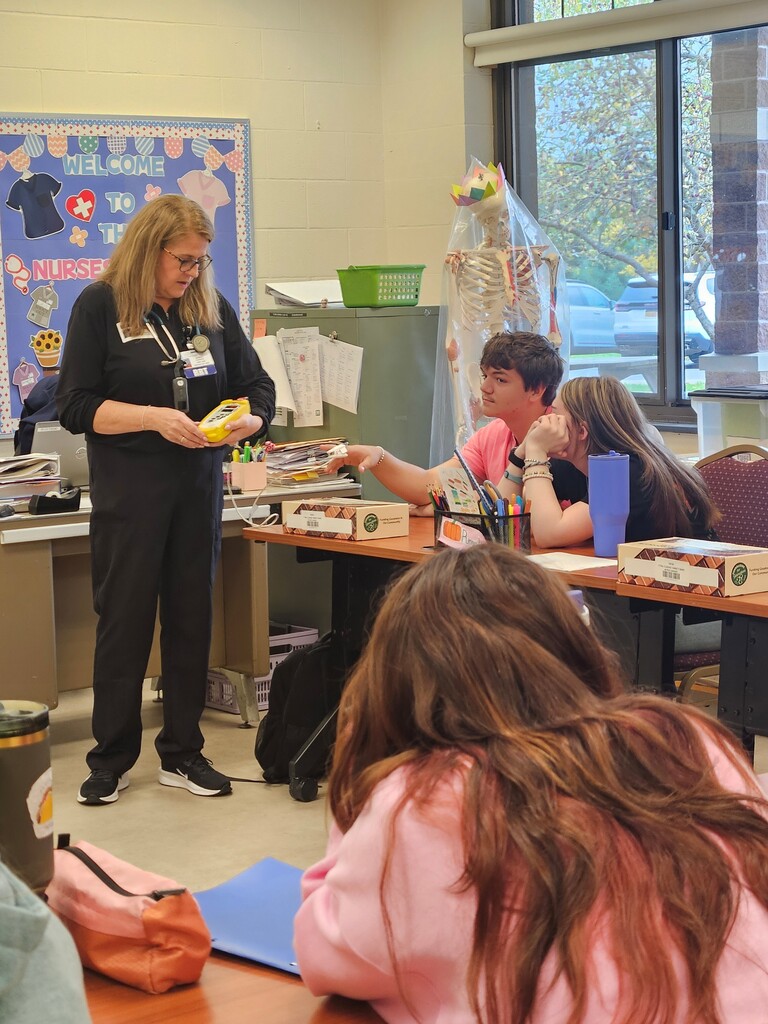 The guest speaker standing in a classroom with a  medical device attached to a student who is sitting at a table. There are other students sitting at tables and watching the presenter.