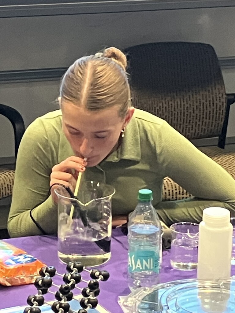 Student sitting at a table with beakers blowing into a straw with a liquid in a beaker.