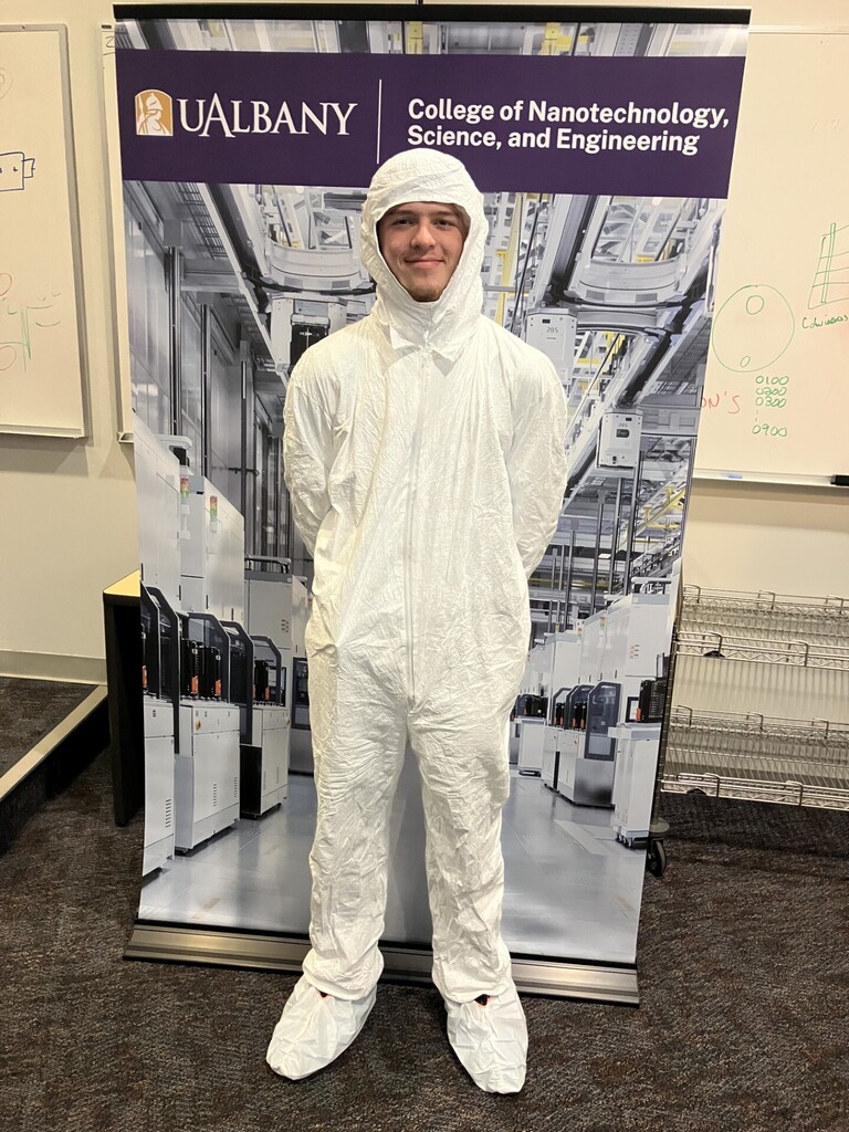Student in white protective suit standing in front of a large UAlbany banner