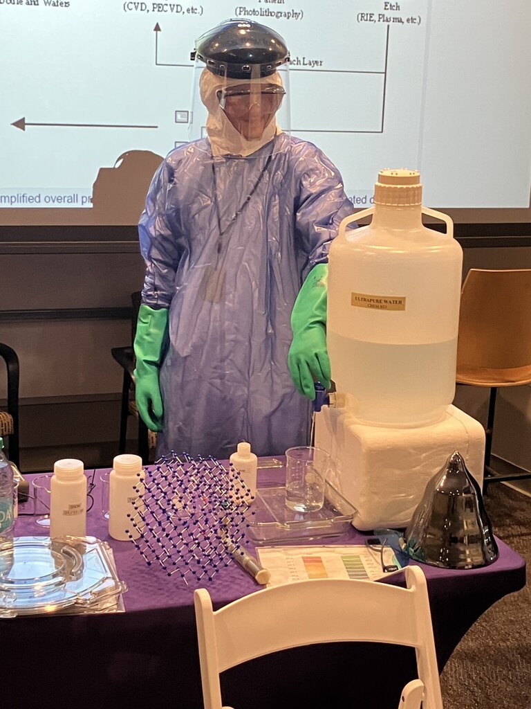 Student in protective gear including green gloves and a face shield. They are standing behind a table with chemical on it.