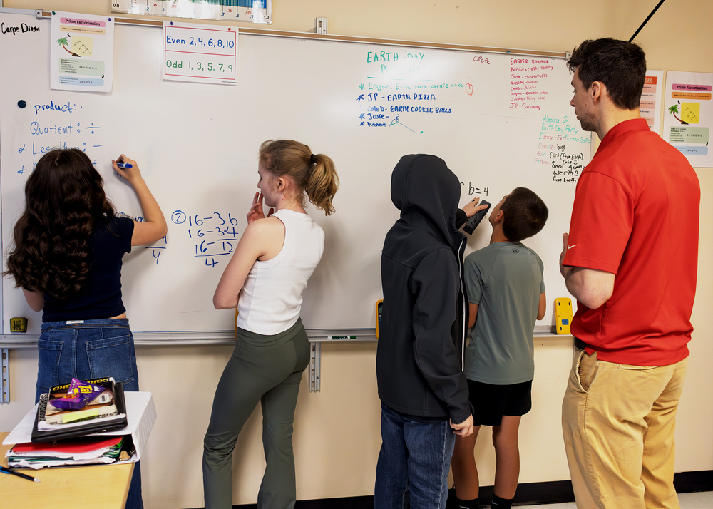 Students work in groups at classroom whiteboards while the teacher observes.