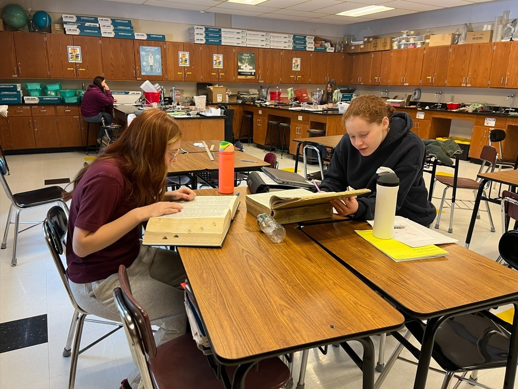 Two students sit at desks in a classroom reading from large hardcover dictionaries while working on an assignment. Additional students can be seen in the background at lab tables. The classroom contains wooden cabinets and science materials along the back wall.