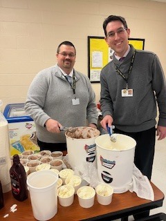 Serving ice cream from large tubs on a table with cups and toppings during a school incentive event.