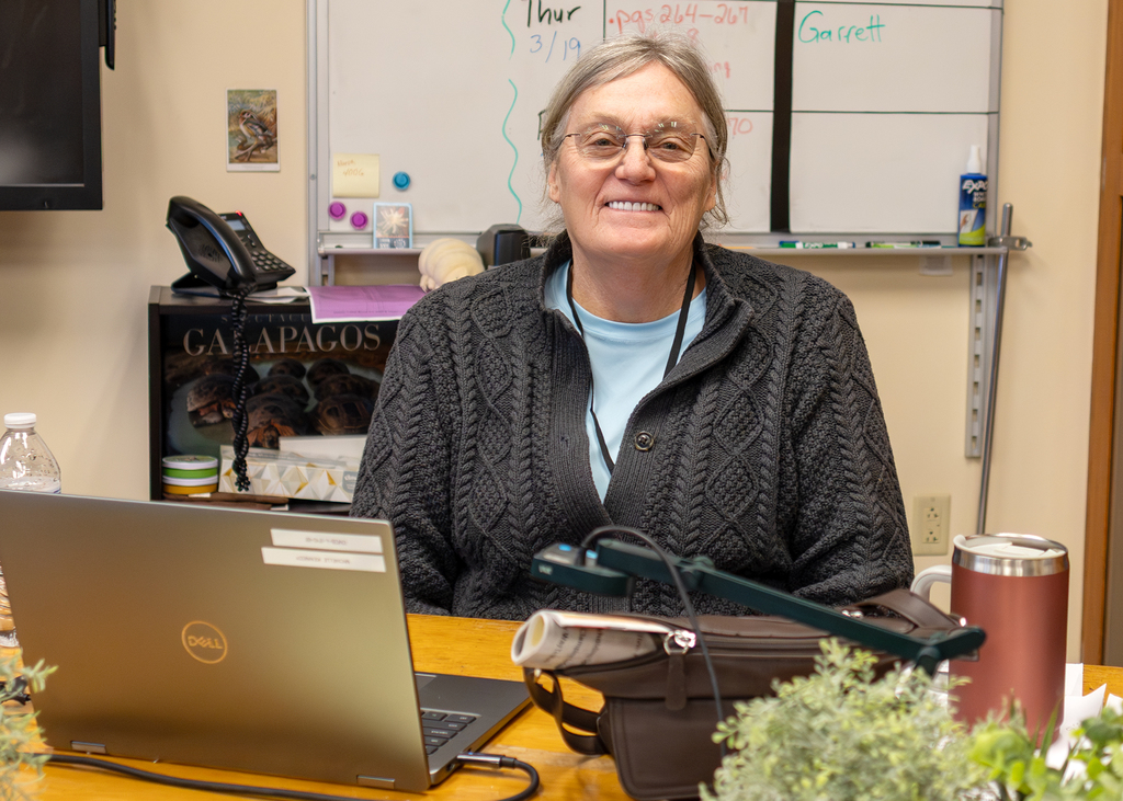 Michelle Kennedy, long-term substitute science teacher at Otselic Valley Central School, seated at a desk in her classroom with a laptop and teaching materials, smiling at the camera.