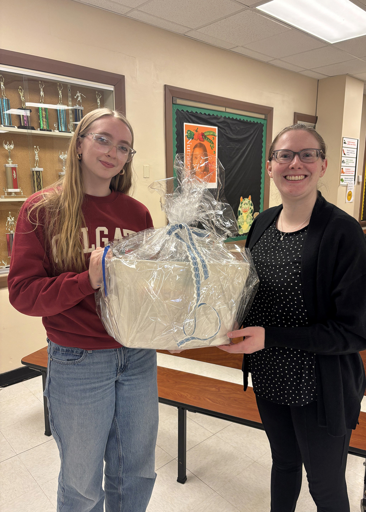 Two women stand indoors holding a wrapped gift basket together and smiling for the camera. The basket is decorated in clear cellophane with blue ribbon. Trophy cases and school hallway displays are visible in the background.