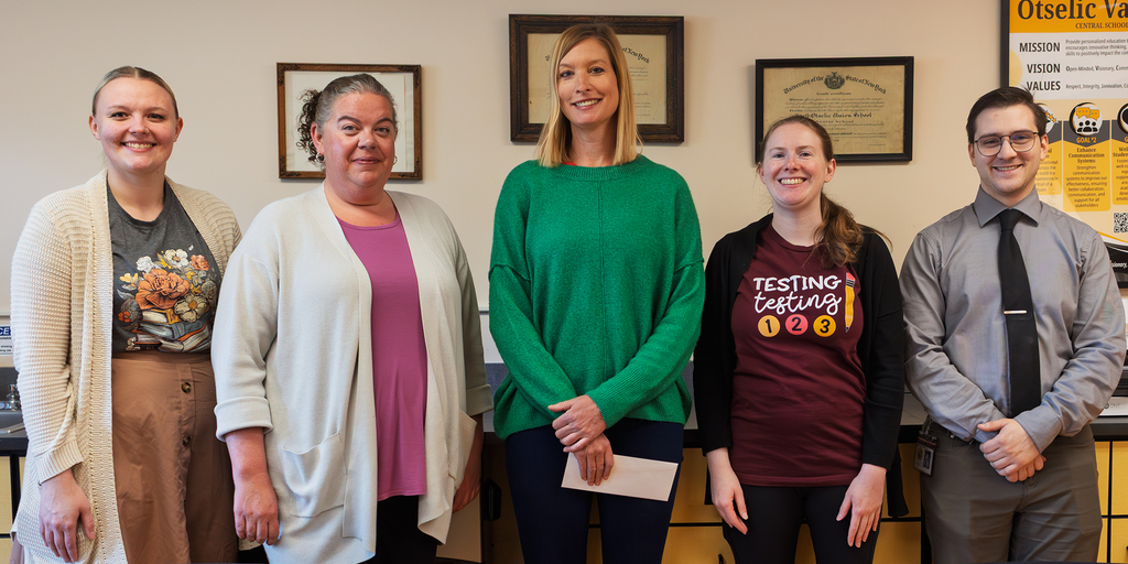 Five Otselic Valley educators stand side by side indoors and smile for a group photo after being recognized for receiving tenure. Framed certificates and a district mission poster are displayed on the wall behind them.