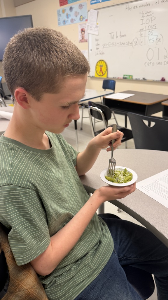 Student tastes ramp pesto pasta while seated at classroom table.