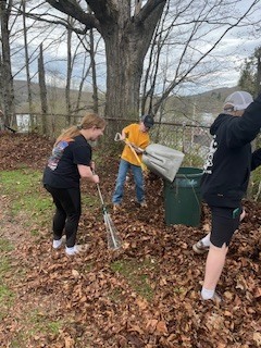 Students rake leaves and clean up around a trash bin near a wooded edge of the cemetery.