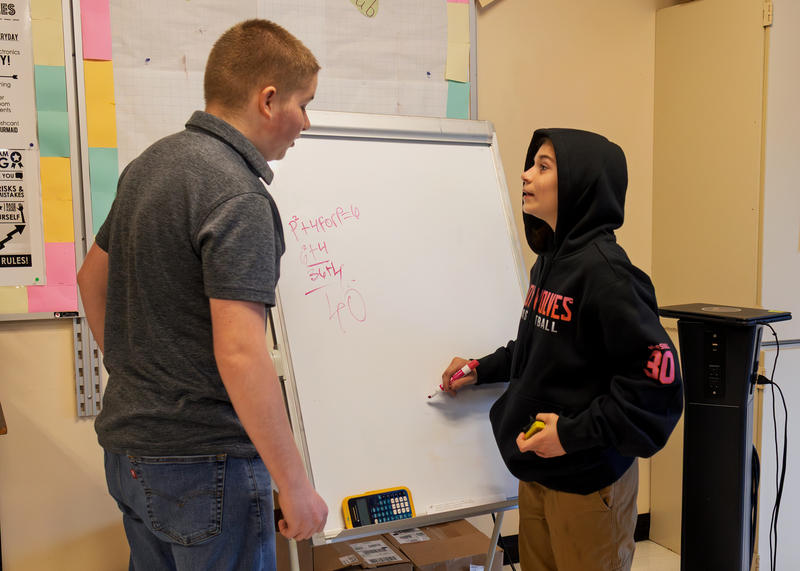 Two students discuss a math problem while writing on a portable whiteboard.