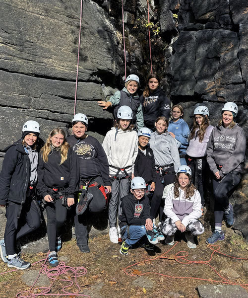 students and chaperones wearing safety gear stand in front of a rock wall with climbing ropes in the background