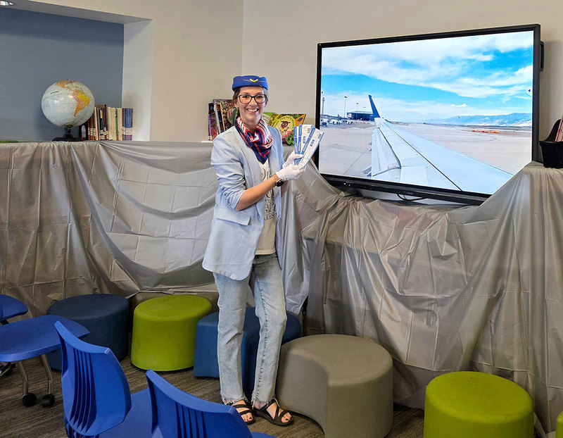 teacher dressed as flight attendant holds airplane tickets while standing in front of screen with image of airport runway