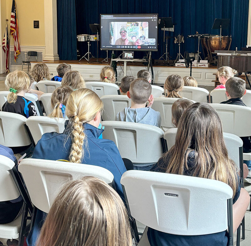 students seated in auditorium  watch virtual visit with author who appears on screen at the front of the room