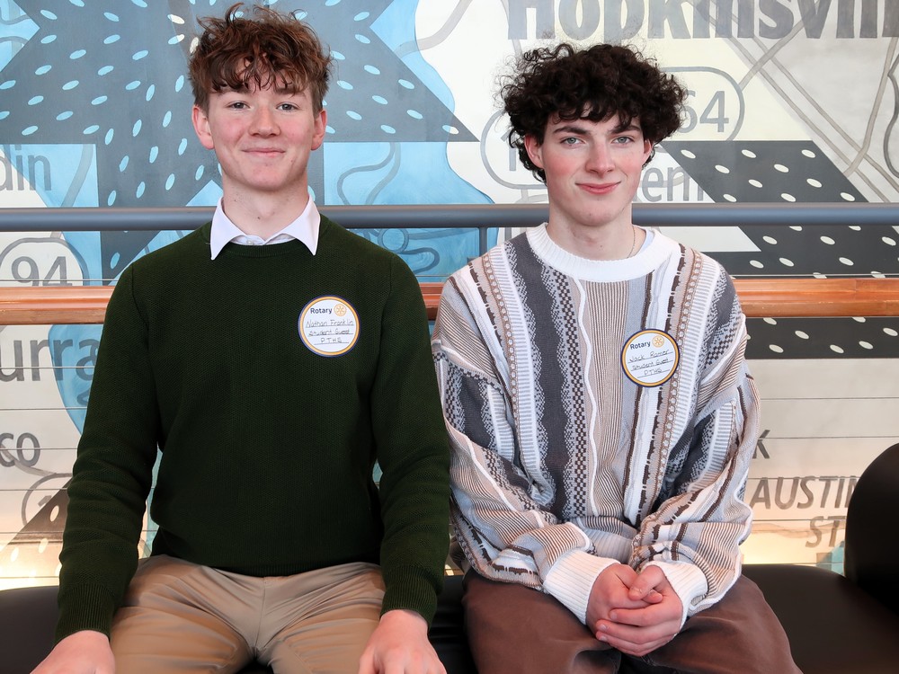 Two students sit on a bench in front of a quilt background