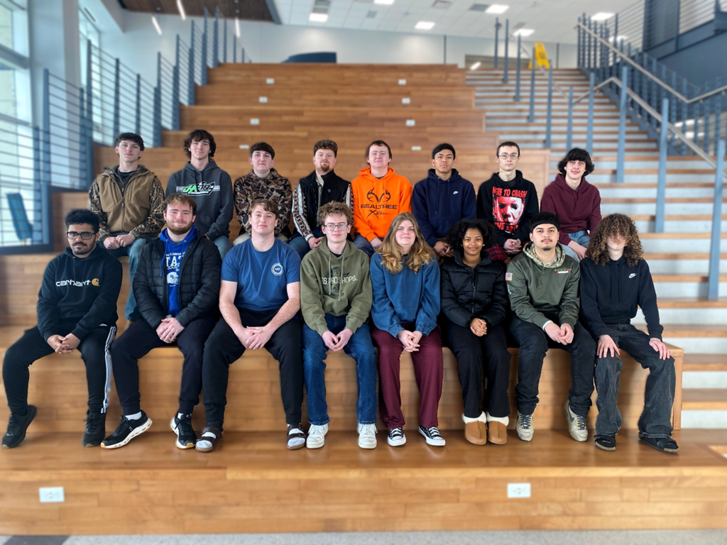 A group of students, seated in a multi-level wooden seating area, smiling and posing for a photo.