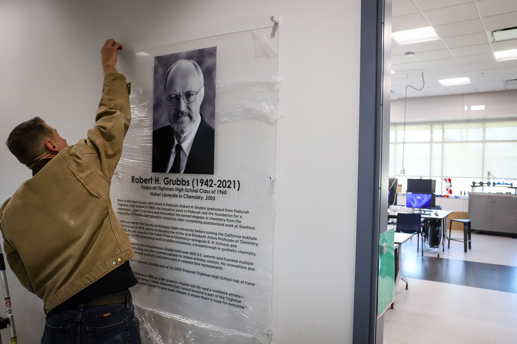 Person in a leather jacket affixing a poster with a black-and-white portrait of an older man.