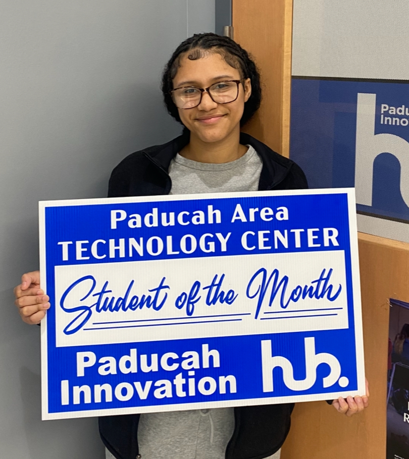 Person in glasses holding blue sign with "Paducah Area Technology Center Student of the Month" text.