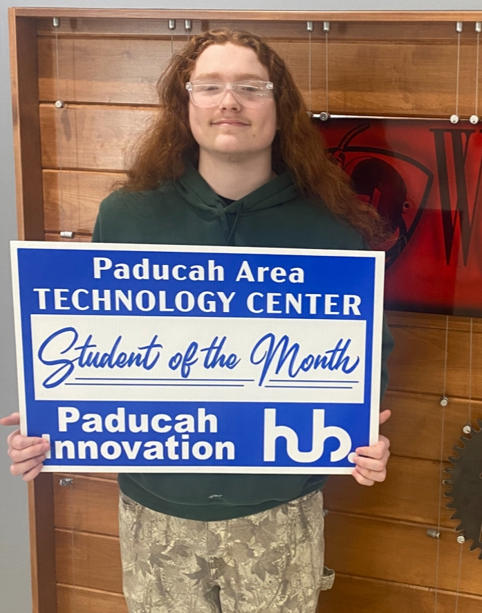 A person in a green hoodie holding a blue plaque that reads "Paducah Area Technology Center Student of the Month".