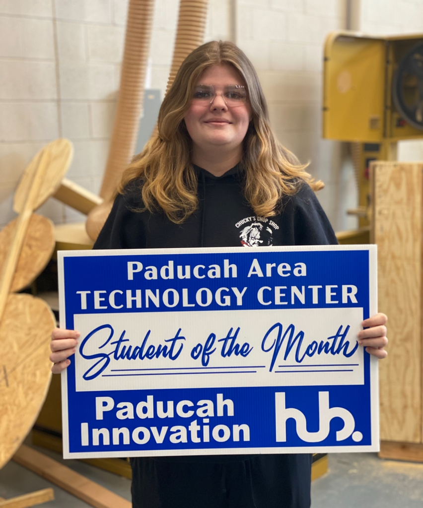 A woman holds a blue sign that reads "Paducah Area Technology Center Student of the Month" in a workshop.