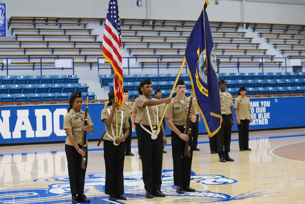 Group of people in uniform standing on a basketball court, holding flags. Empty blue bleachers in the background.