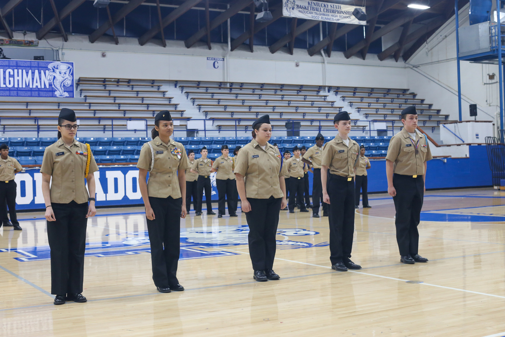 Seven uniformed cadets stand on a basketball court, facing forward. Empty bleachers are in the background.