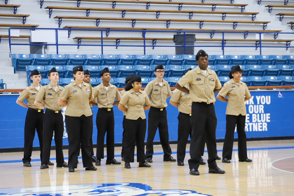 A group of uniformed individuals, possibly cadets, stands in rows on a basketball court with empty bleachers.