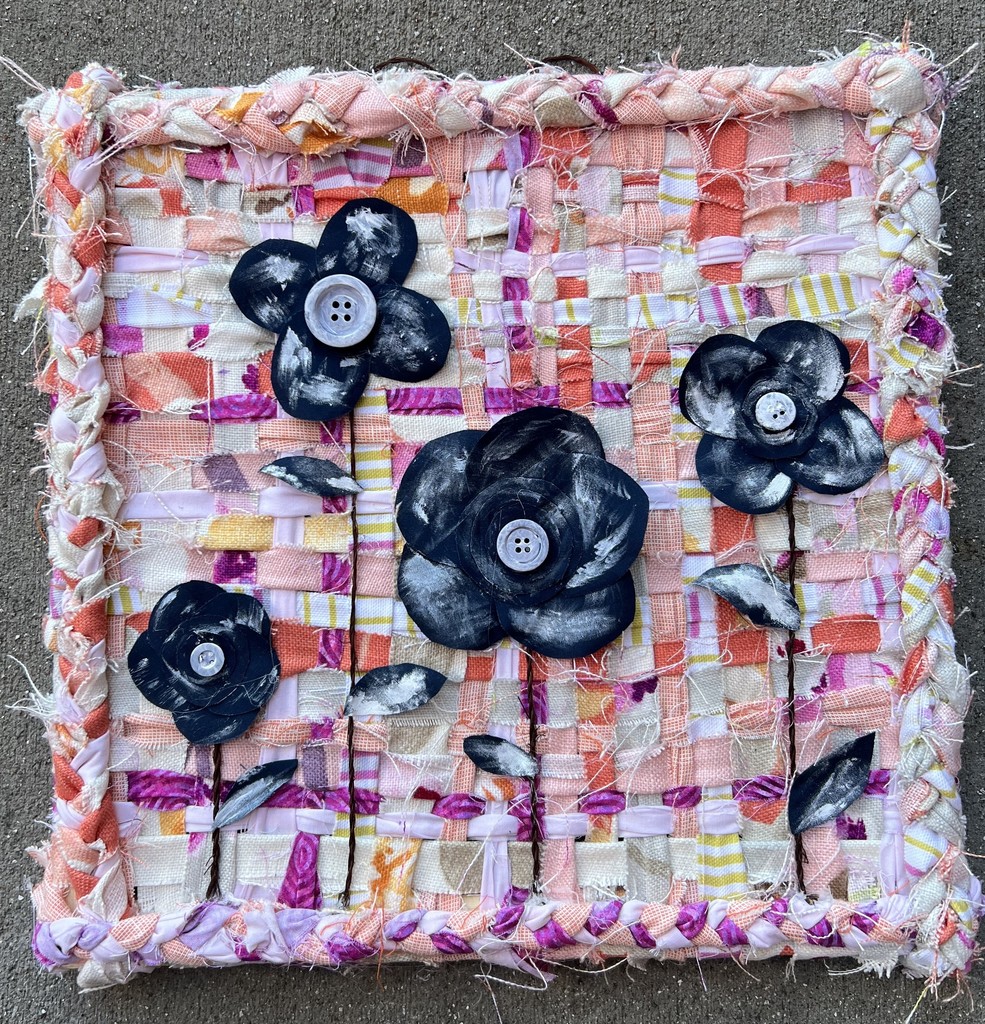 A square, colorful fabric with flowers and buttons. It has a pink border and a textured, layered appearance.