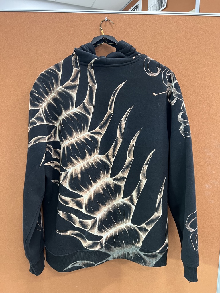 A black hoodie with a white flame design hangs on a brown wall. The design resembles a flame with sharp points.