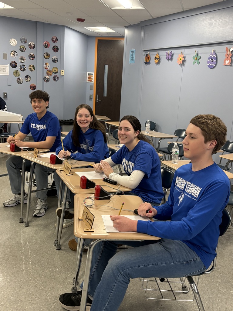 Four students in blue shirts sit at desks in a classroom. Papers, pens, and red cups are on the desks.