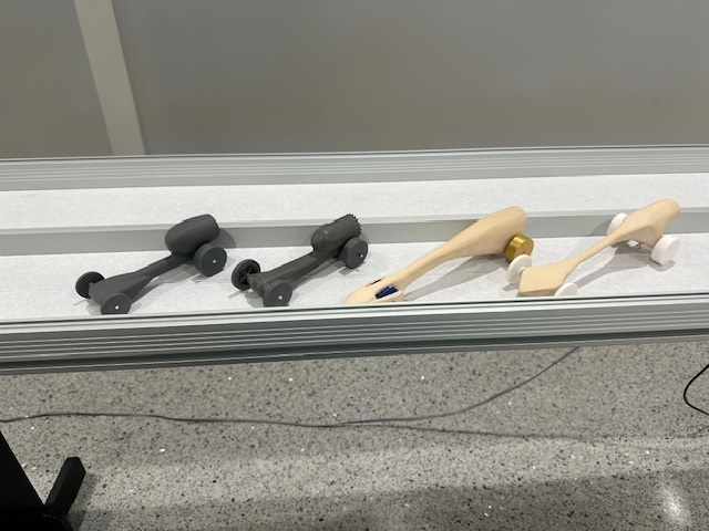 Four small vehicles, three black and one light brown, on a white shelf. The black vehicles have wheels, and the brown vehicle has a yellow ball.