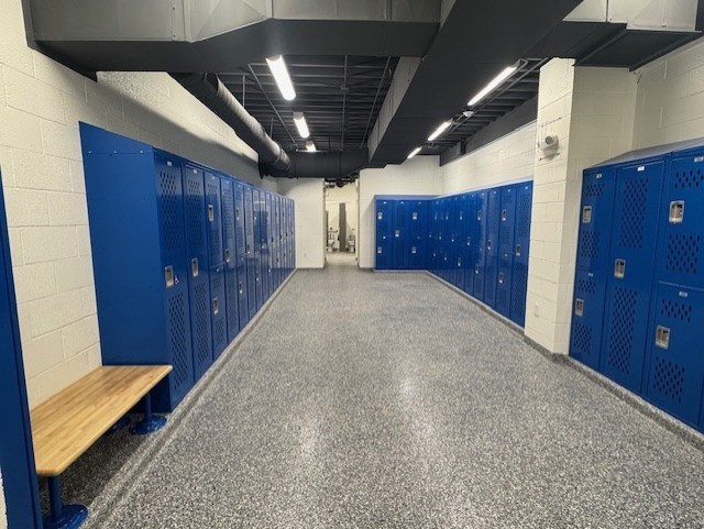 Empty locker room with rows of blue lockers and a wooden bench. The room has gray floors and white walls.